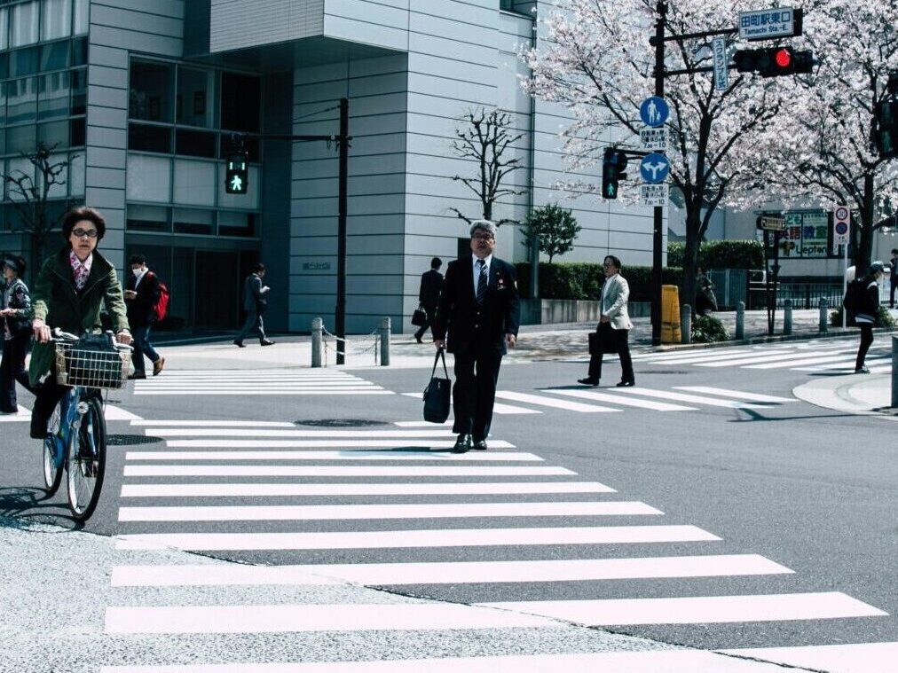 Pedestrians crossing a busy intersection in Nagawa, Japan with cherry blossoms and modern architecture.